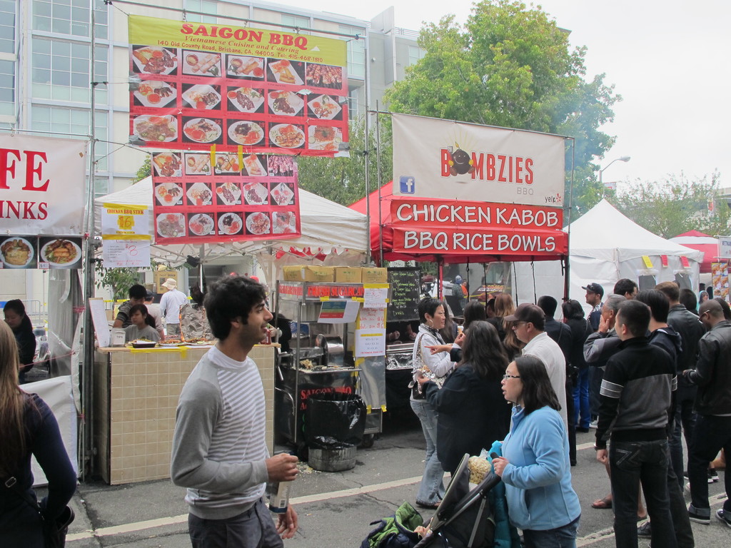 Menschen stehen auf einer Straße außerhalb der Stadt, wobei ein Mann in einem grauen und weißen T-Shirt eine Wasserflasche hält und eine Frau in einer blauen Jacke einen Kinderwagen schiebt. Im Hintergrund sind Stände mit weißen und roten Zelten und bunten Bannern sowie Gebäude, Bäume und eine Straßenlaterne zu sehen.