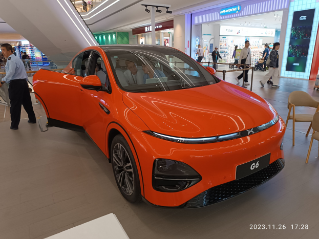 Oranges Elektroauto in einer Ausstellungshalle mit einer Gruppe von Menschen drumherum, Stühlen und einem Wagen auf der rechten Seite und Geschäften und Namensschildern im Hintergrund.