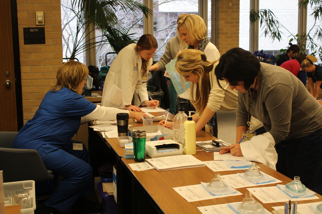 Ein Raum mit Menschen, die an einem Schreibtisch stehen, eine Frau in einem blauen Kleid in der Ecke sitzt, Papiere, Bücher und Flaschen auf dem Schreibtisch, große Glasfenster und Pflanzen im Raum.