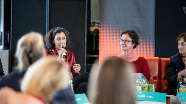 Eine Gruppe von Frauen sitzt in der ersten Reihe, wobei eine links eine rote Kleidung trägt und ein Mikrofon hält, und im Hintergrund eine Laufbandrolle und ein schwarzes Tuch zu sehen sind.