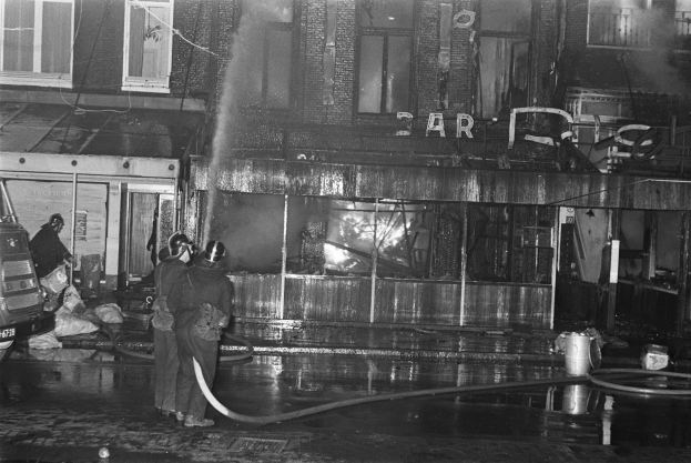 Feuerwehrleute bei der Brandbekämpfung an einer Straßenecke, mit Menschen mit Rohren, einem geparkten Fahrzeug, verstreuten Gegenständen, einem Gebäude mit Fenstern, einem Schild und Drähten sowie einem bewölkten Himmel.