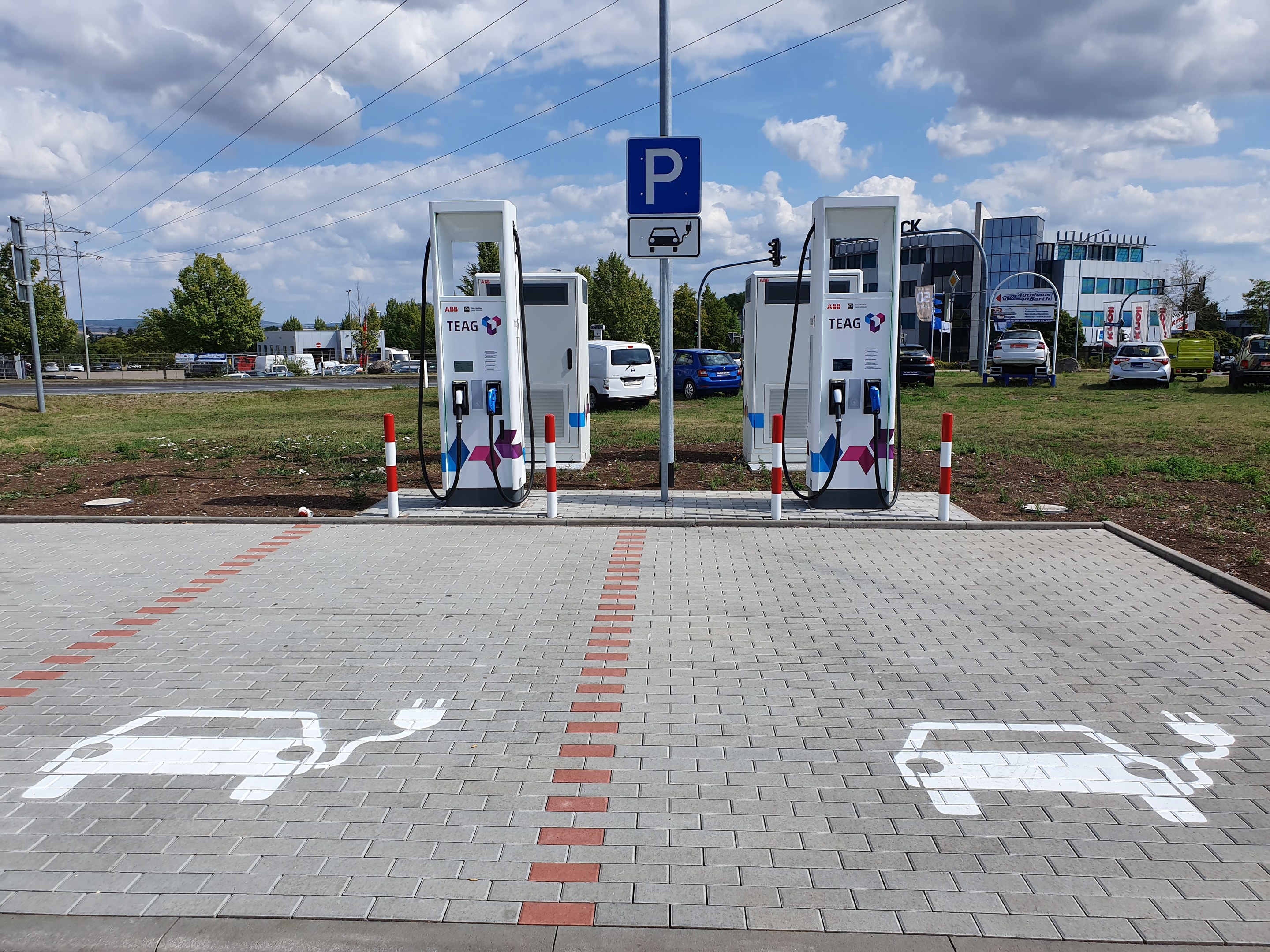 Elektroauto-Ladestation auf einem Parkplatz mit Gebäuden, Bäumen, Strommasten und Fahrzeugen auf der Straße unter einem bewölktem Himmel.