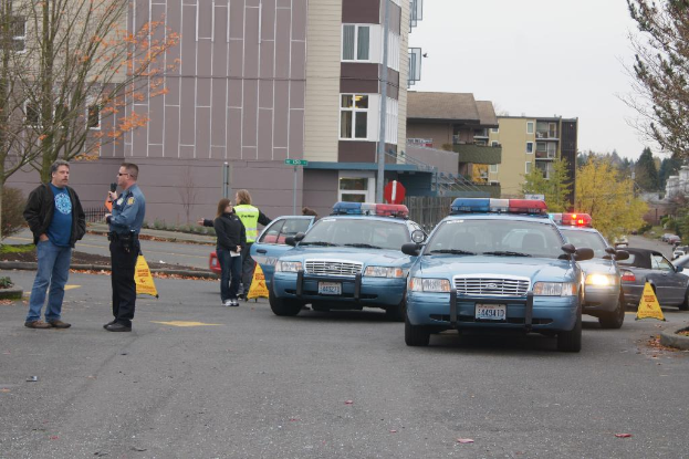 Autos auf einer Straße mit vier Menschen in der Nähe, Gebäude mit Fenstern im Hintergrund, Bäume und Warnkegel.