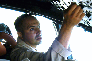 Ein Mann in einem blauen Hemd und schwarzer Mütze sitzt im Fahrersitz eines hellblauen Autos und hält das Lenkrad fest mit beiden Händen, wobei er den Eindruck eines Taxifahrers mit getönten Scheiben macht.