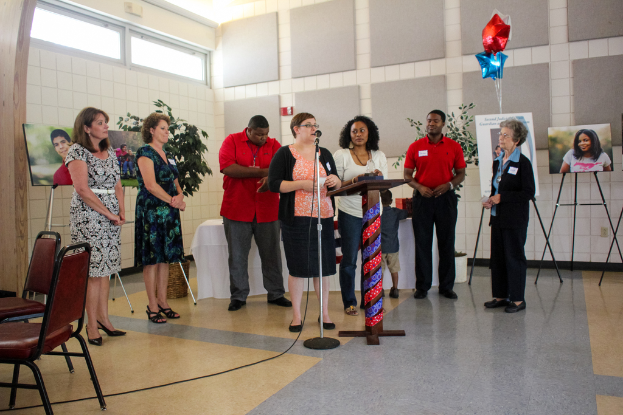 Eine Frau steht an einem Podium mit einem Mikrofon und spricht in einem Raum mit zwei leeren Stühlen, einem Tisch mit einer weißen Decke, Topfpflanzen, einem roten und blauen sternförmigen Ballon und geschlossenen Fenstern.