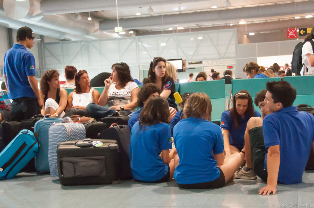 Ein viel frequentierter Flughafen-Terminal mit vielen Menschen, die auf Fl√ľge warten, Gep√§ck auf der linken Seite und Glast√ľren mit Richtungsschildern im Hintergrund.