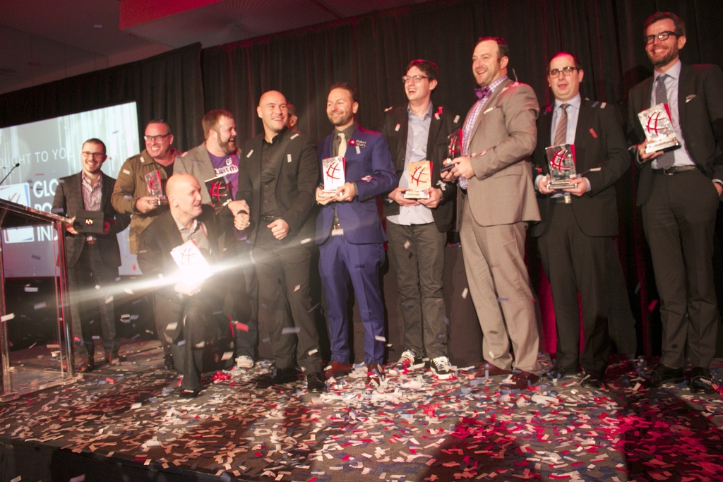 Eine Gruppe von Jungs steht auf einer Bühne und hält Schilde, mit einem Mann in der Mitte, der Licht reflektiert, einem Podium mit einem Mikrofon links daneben, einem Bildschirm und einem schwarzen Vorhang im Hintergrund sowie verstreuten Papierstücken auf der Bühne.