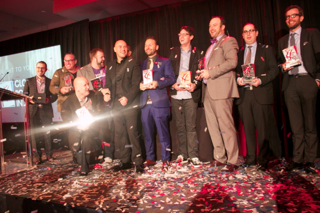 Eine Gruppe von Jungs steht auf einer Bühne und hält Schilde, mit einem Mann in der Mitte, der Licht reflektiert, einem Podium mit einem Mikrofon links daneben, einem Bildschirm und einem schwarzen Vorhang im Hintergrund sowie verstreuten Papierstücken auf der Bühne.