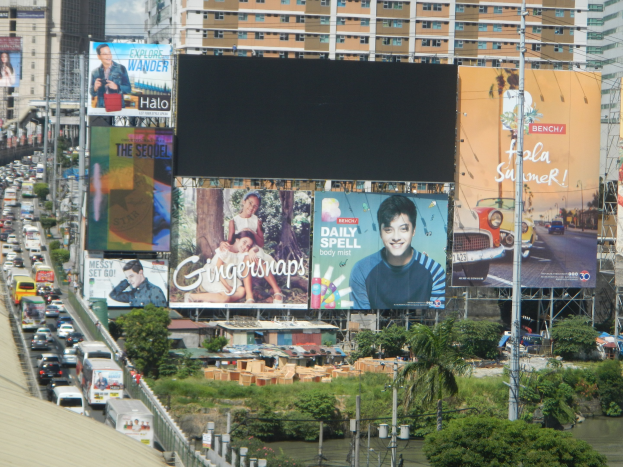 Stadtstraße mit starkem Verkehr, hohen Gebäuden, einem zentralen Werbeplakat, Bäumen, Pflanzen, Gras, Wasser, Strommasten mit Drähten und einem bewölkten Himmel.