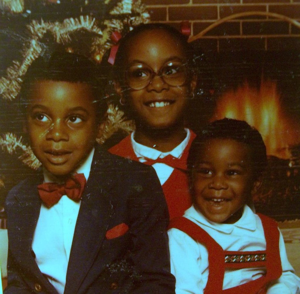 Drei lachende Kinder vor einem Weihnachtsbaum mit einem Kamin aus Holz auf der rechten Seite.