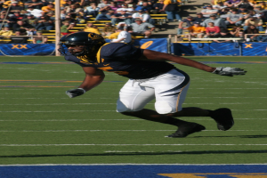 Ein Fußballspieler in voller Montur rennt mit dem Ball auf einem Feld, während Zuschauer auf Stühlen, Geländern, Treppen und einem Pfahl im Hintergrund sitzen, mit sichtbaren Bannern.