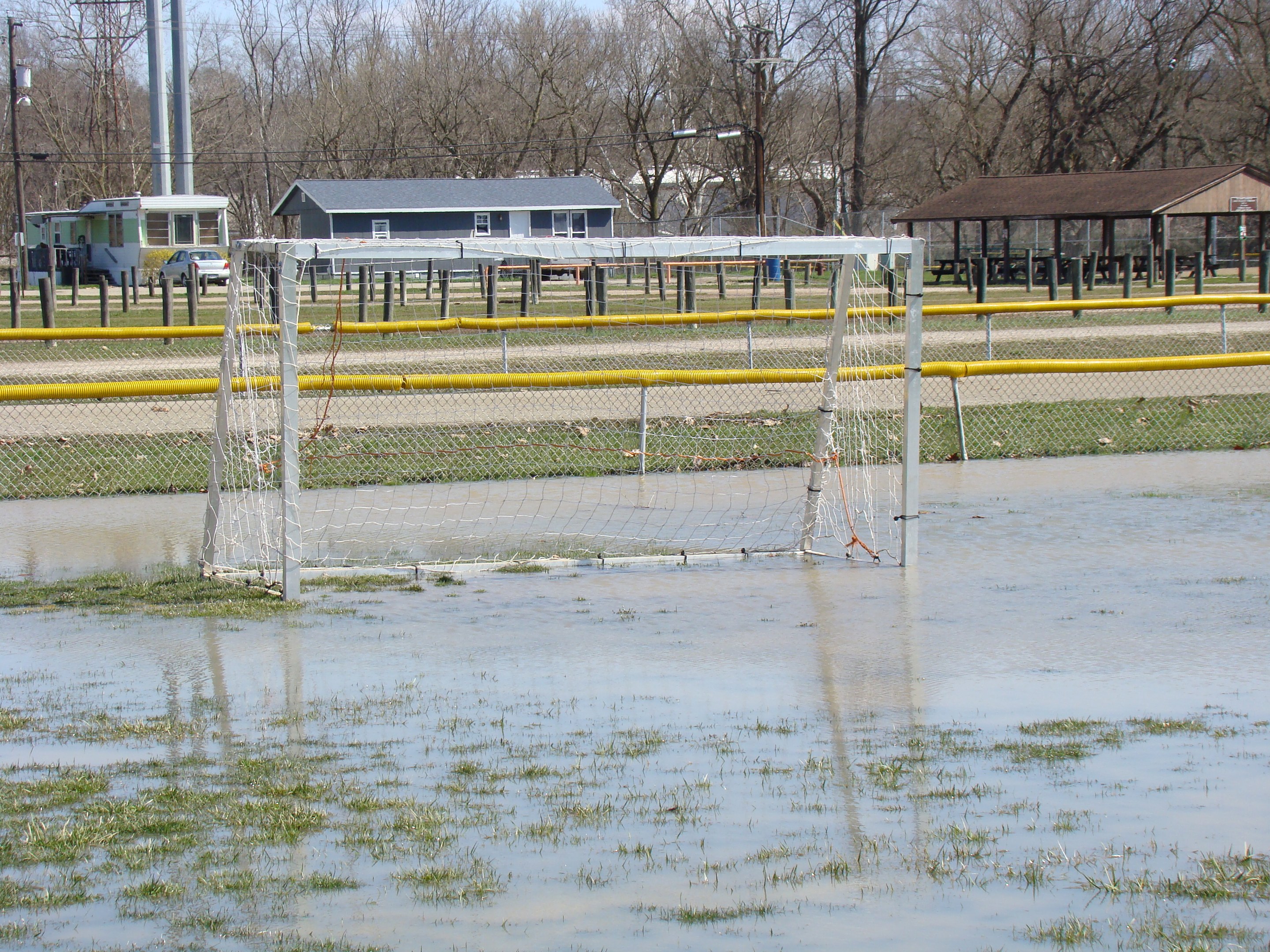 Ein Fußballtor steht in einem überfluteten Feld mit Gras und Wasser, umgeben von Hütten, Pfählen, Bäumen, Fahrzeugen und einem klaren blauen Himmel im Hintergrund.