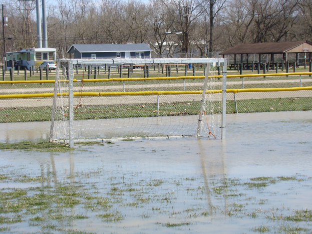 Ein Fußballtor steht in einem überfluteten Feld mit Gras und Wasser, umgeben von Hütten, Pfählen, Bäumen, Fahrzeugen und einem klaren blauen Himmel im Hintergrund.