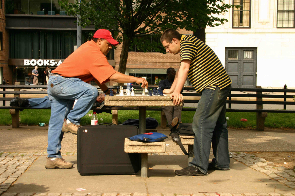 Eine Stadtansicht mit einem Tisch und Bänken, einem Schachbrett mit Stücken auf dem Tisch, Taschen auf den Bänken, einem Mann in rotem Hut mit einem großen Koffer, einem anderen schlafenden Mann auf einer Bank und einem Gebäude mit Fenstern, Türen, einem Baum und Gras im Hintergrund.