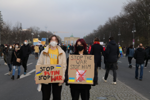 Eine Gruppe von Menschen in Masken, die eine Straße in Berlin entlanggehen und "Stop the War"-Schilder halten, mit Bäumen, Laternenpfählen, einem Bogen und einem klaren blauen Himmel im Hintergrund.