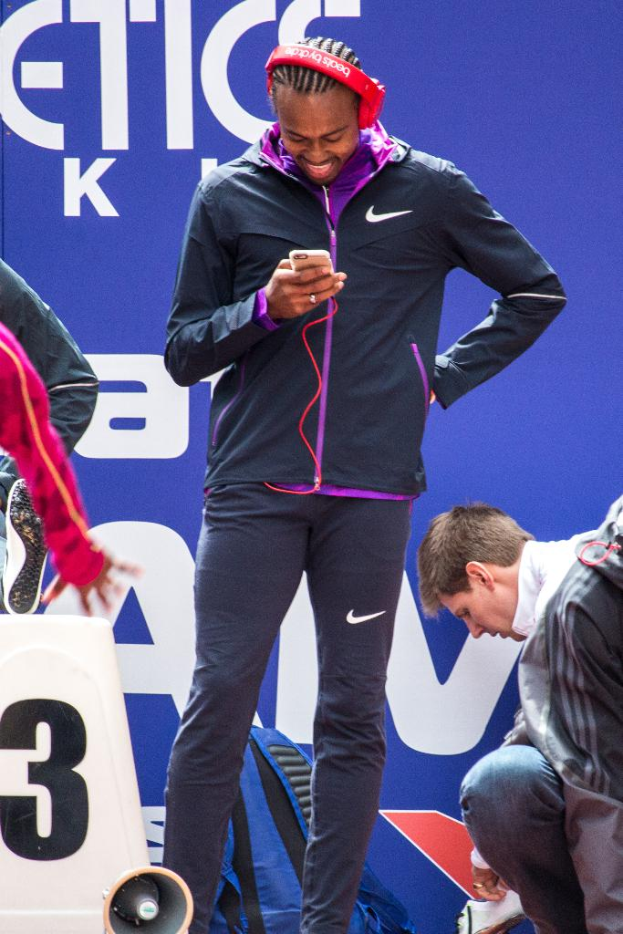 Eine Person, die mit einem Mobiltelefon in der Hand steht und lächelt, daneben eine weitere Person in Hockstellung, ein Rucksack, ein Megafon unten, eine Wand im Hintergrund und eine weitere Person, die steht.