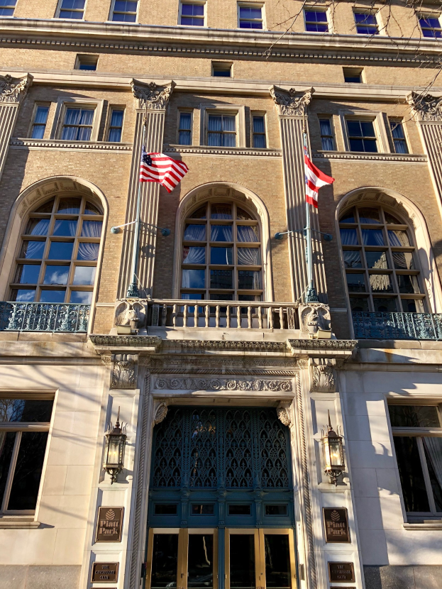 Außenansicht des Plaza Hotels in New York City, mit Glasfenstern, Türen, Säulen, Geländern, Lampen, Texttafeln und Flaggen vor einem Himmelhintergrund.