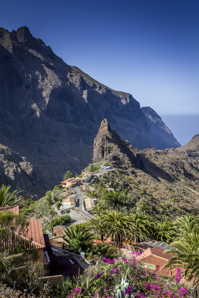Eine ländliche Szene mit Häusern, Bäumen und Felsen im Vordergrund, einem Hügel im Mittelgrund und dem Himmel im Hintergrund.