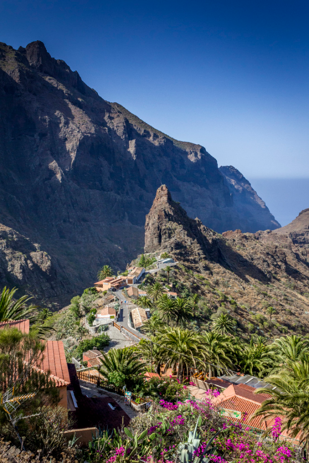 Eine ländliche Szene mit Häusern, Bäumen und Felsen im Vordergrund, einem Hügel im Mittelgrund und dem Himmel im Hintergrund.