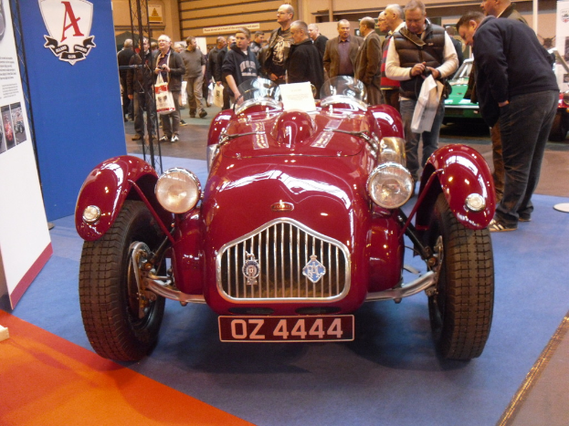 Ein Fahrzeug auf einer Ausstellung mit zwei Bannern auf der linken Seite, umgeben von Menschen, anderen Fahrzeugen im Hintergrund auf der rechten Seite und einer Wand dahinter.