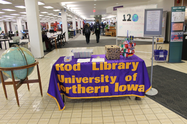 Eine Ausstellung mit einem Tisch, auf dem verschiedene Landesflaggen, eine Box, Papiere und ein Banner liegen, eine Kugel links daneben, eine Anschlagtafel rechts daneben mit ein paar Leuten in der Nähe und zahlreichen Tischen und Stühlen im Hintergrund.