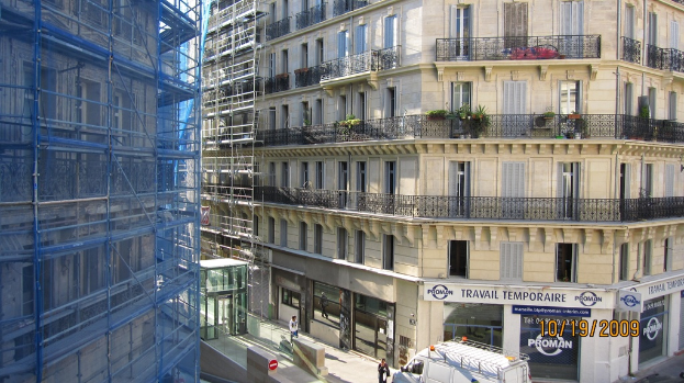 Ein Bus steht auf einer Straße mit einer Wand, Strommasten mit Kabeln, Bäumen, Gebäuden und dem Himmel im Hintergrund.