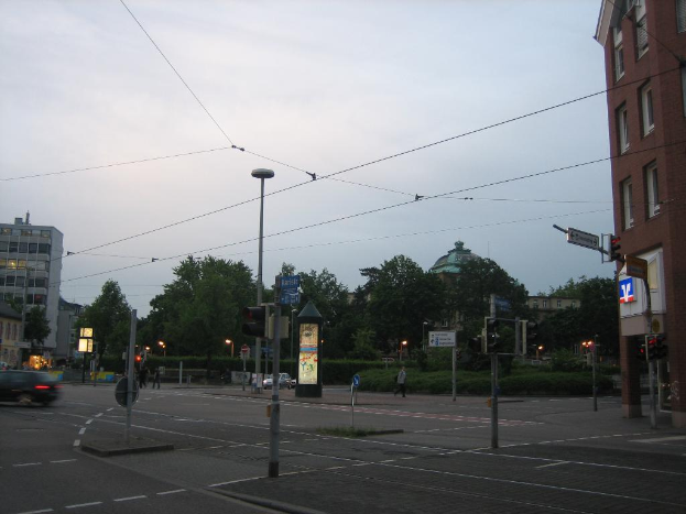 Masten mit Verkehrszeichen und Schildern säumen einen Weg mit Fahrzeugen und Menschen, mit Bäumen, Gebäuden, Kabeln und Himmel auf der rechten Seite.