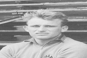 Schwarz-weißes Foto eines Mannes in einer Jacke, der vor gestapelten Holzplanken steht, der als legendärer deutscher Fußballspieler gilt.