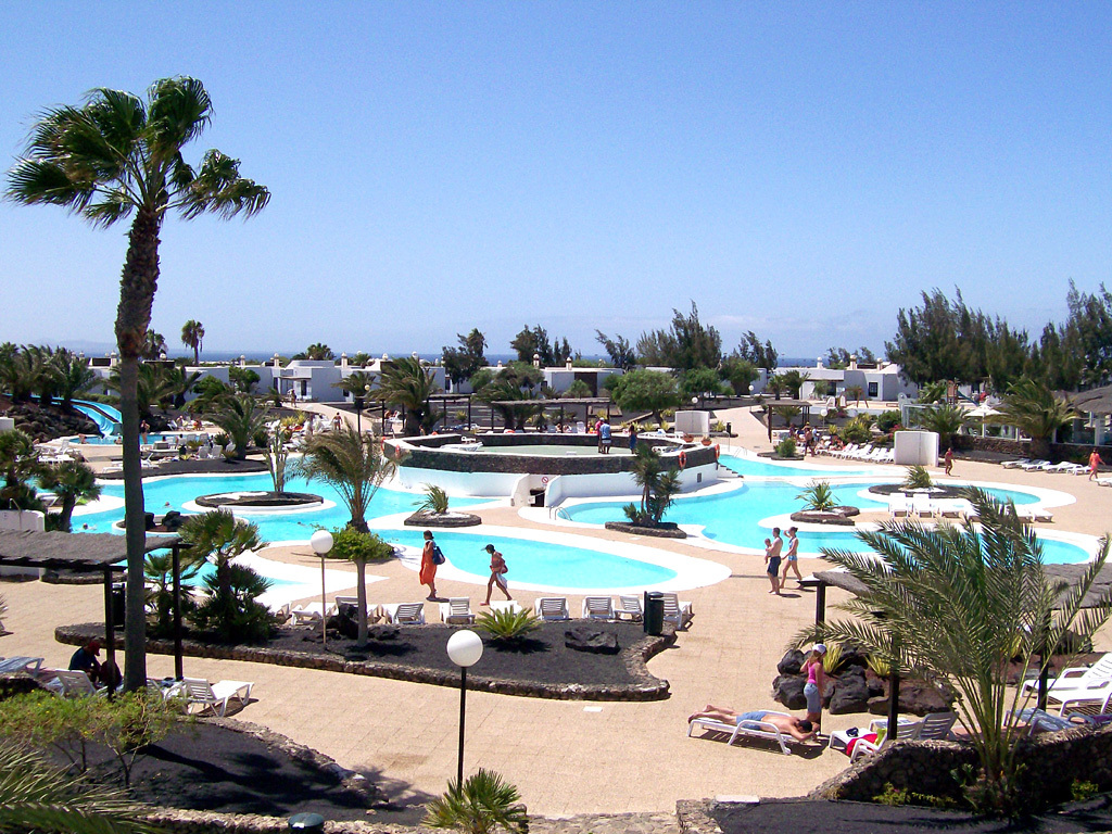 Ein Resort mit Pools umgeben von Bäumen, Menschen zwischen ihnen, Gebäuden im Hintergrund und einem klaren Himmel darüber.