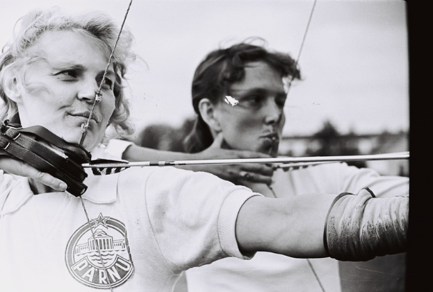 Zwei Frauen stehen nebeneinander, jede hält einen Bogen und einen Pfeil in der Hand.