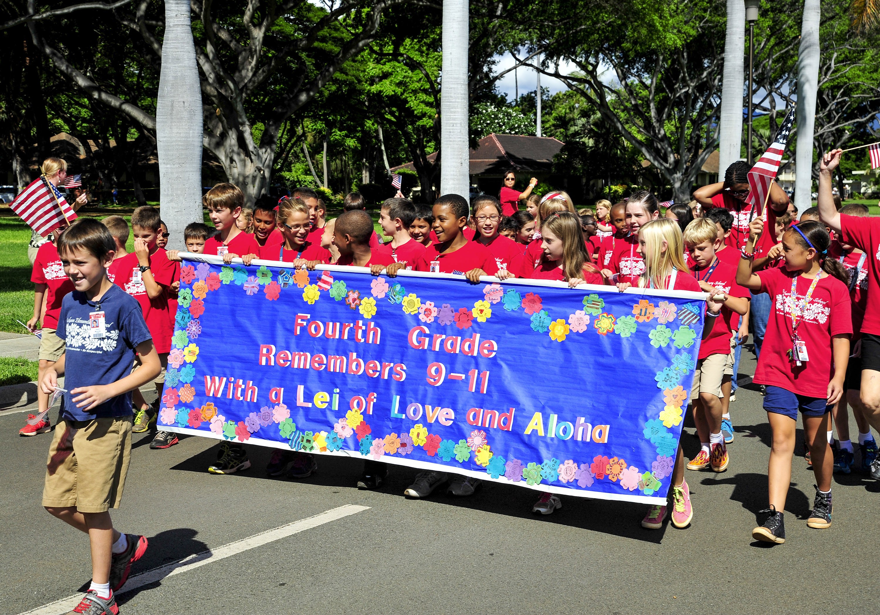 Gruppe von Kindern in roten T-Shirts auf einer Straße marschierend, mit einer Fahne und Flaggen, mit Bäumen und Häusern im Hintergrund.