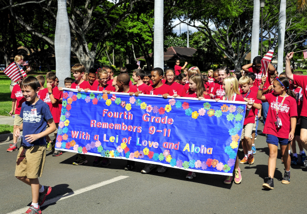 Gruppe von Kindern in roten T-Shirts auf einer Straße marschierend, mit einer Fahne und Flaggen, mit Bäumen und Häusern im Hintergrund.