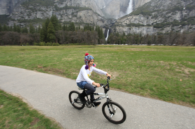 Ein Mädchen fährt Fahrrad mit einem Lächeln im Gesicht, umgeben von Gras, mit Bergen und Bäumen im Hintergrund.