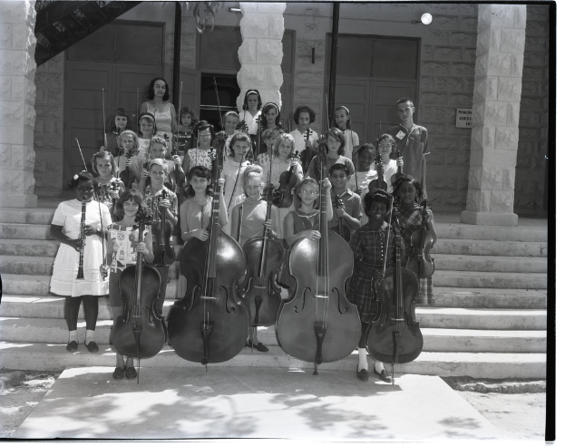 Kinder auf einer Treppe mit Musikinstrumenten wie Celli, Geigen und Trompeten, im Hintergrund Säulen und eine Wand mit Türen.