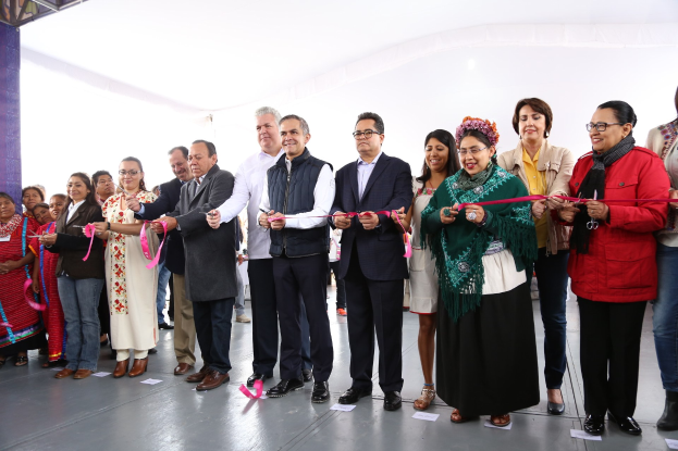 Eine Gruppe von Menschen schneidet bei einer Zeremonie eine Schnur durch, lächelt und hält eine Schere in der Hand vor einer Menge, mit einer Säule auf der linken Seite, verstreuten Papieren auf dem Boden und einem weißen Tuch im Hintergrund.