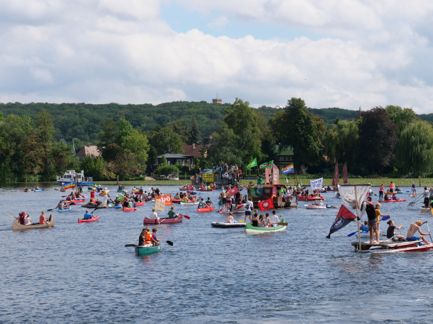 Eine große Gruppe von Menschen in Kanus auf einem Gewässer, einige halten Paddel und Fahnenstangen, mit Bäumen, Gebäuden und einem bewölkten Himmel im Hintergrund während eines Kanurennens.