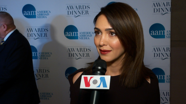 Emmy Rossum in einem schwarzen Kleid mit einem Mikrofon, mit einem Mann zu ihrer Linken und einem Banner mit der Aufschrift "Aam Awards Dinner" im Hintergrund.