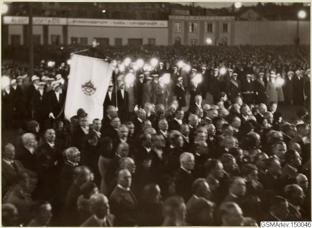 Schwarzes und weißes Foto einer großen Menschenmenge vor einem Gebäude, mit einer Person, die eine Fahne in der Mitte hält, sowie Masten und Lichter im Hintergrund und einem Text unten, der "Gustav Klimts Rede an der Berliner Mauer" lautet.