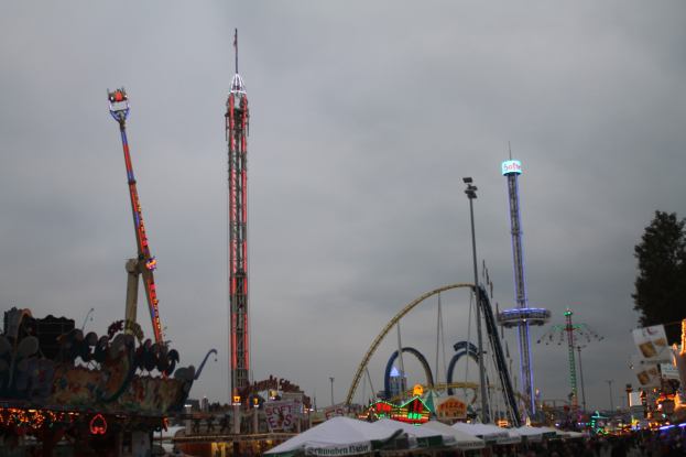 Eine Menschenmenge bei einer nächtlichen Karnevalsveranstaltung, beleuchtet von bunten Lichtern an Fahrgeschäften und Pfählen, mit bewölktem Himmel.