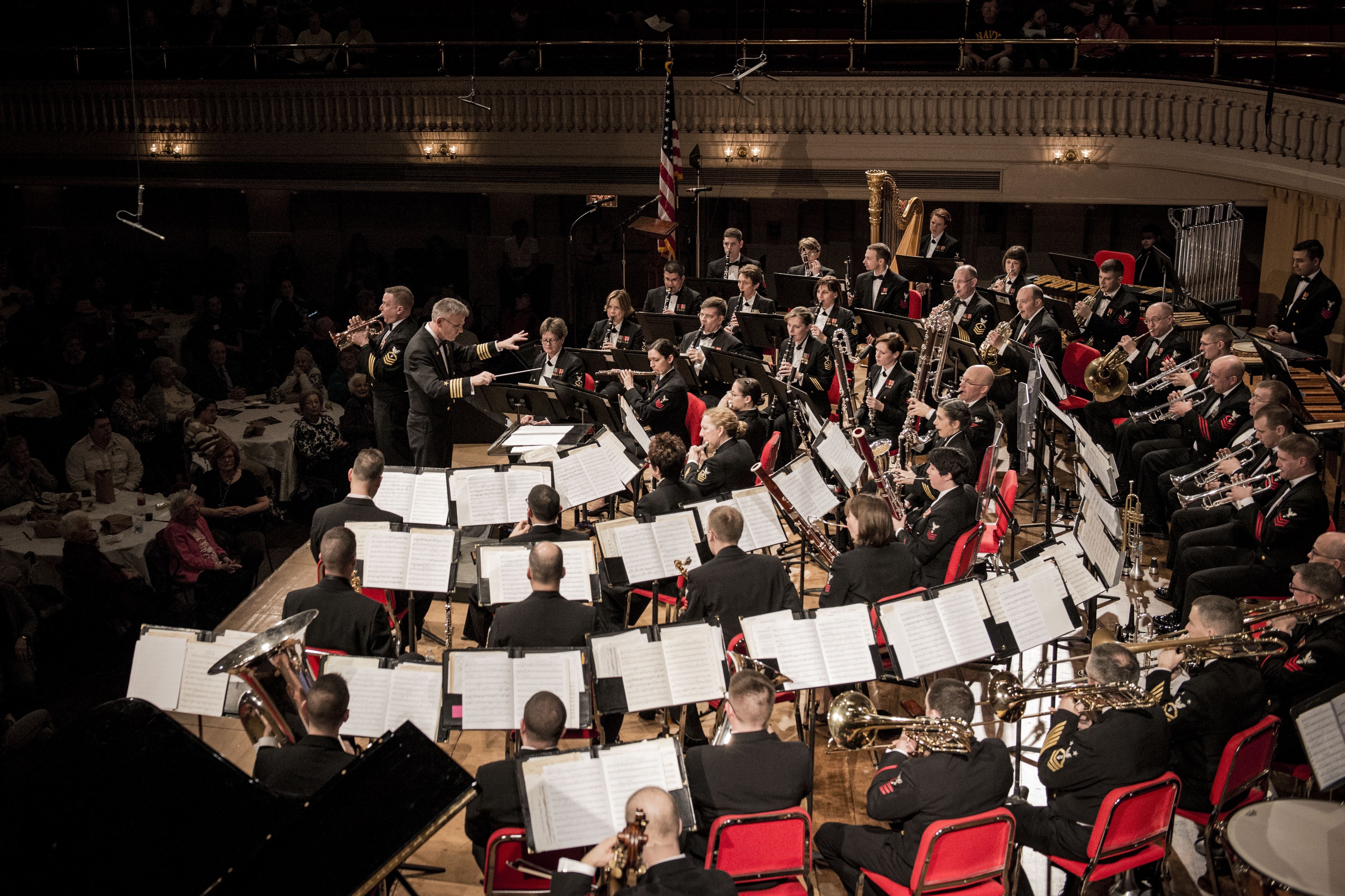 Große Militärmusikformation in einem Konzertsaal mit sitzenden und stehenden Musikern, Notenständern und einer Flagge an der Rückwand.