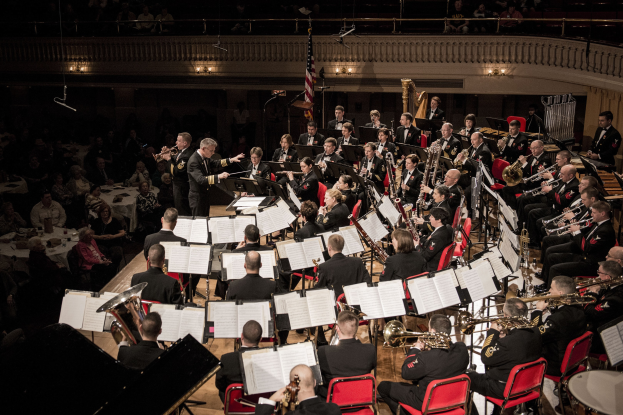 Große Militärmusikformation in einem Konzertsaal mit sitzenden und stehenden Musikern, Notenständern und einer Flagge an der Rückwand.
