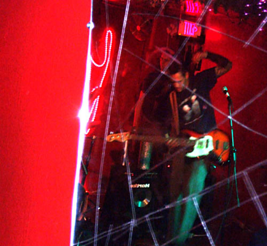 Ein Mann in einem schwarzen T-Shirt steht in der Mitte und hält eine Gitarre, während ein anderer Mann hinter ihm möglicherweise Instrumente spielt, unter bunten Lichtern vor einer roten Wand.