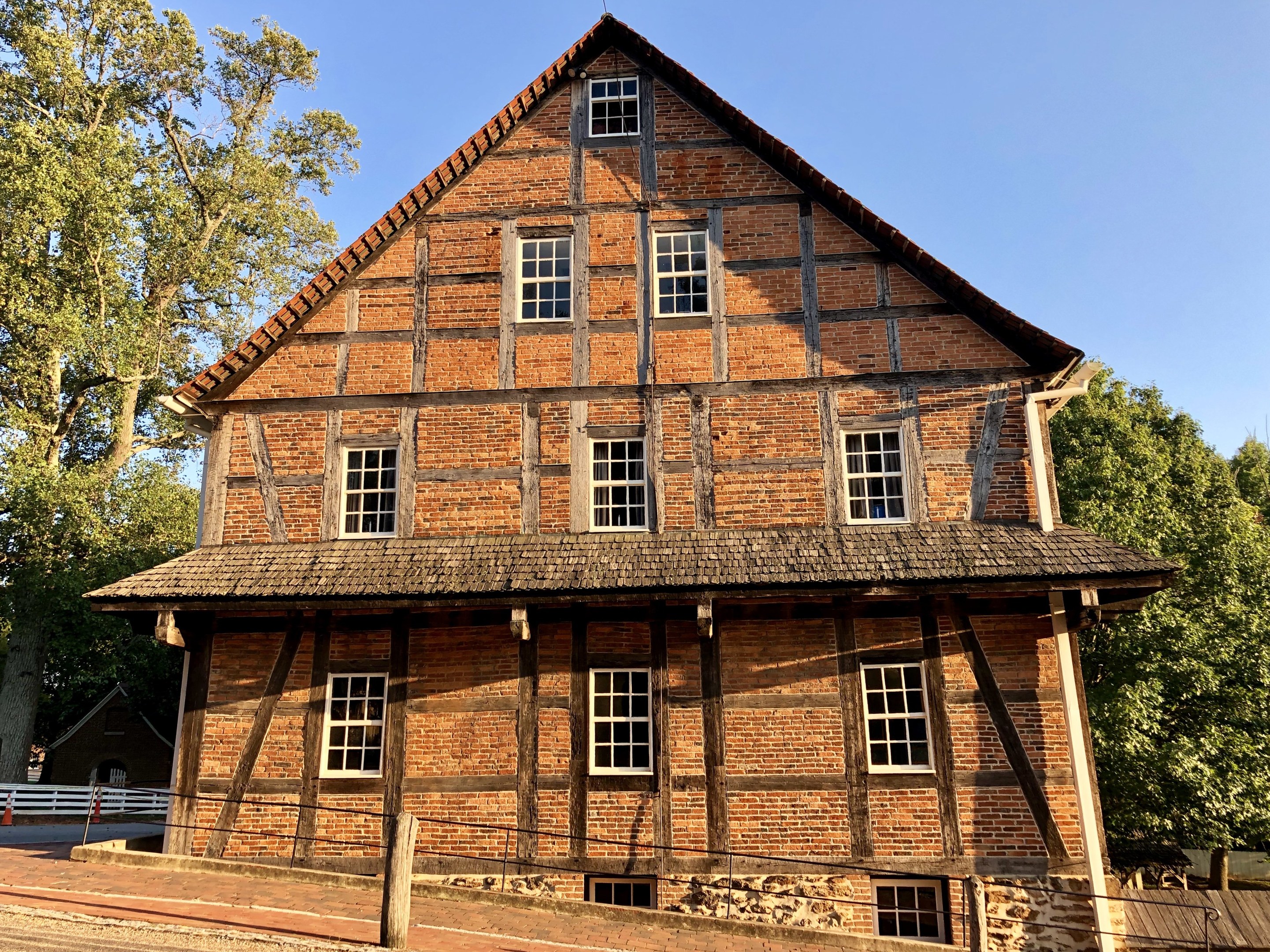 Alter Backsteinbau mit einem zentralen Uhrenturm, umgeben von einem Zaun und einer Straße, vor einem Wald und einem klaren blauen Himmel.