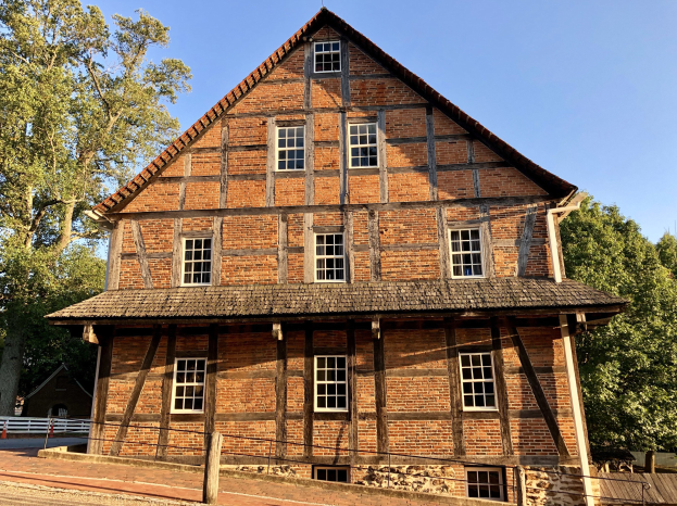 Alter Backsteinbau mit einem zentralen Uhrenturm, umgeben von einem Zaun und einer Straße, vor einem Wald und einem klaren blauen Himmel.