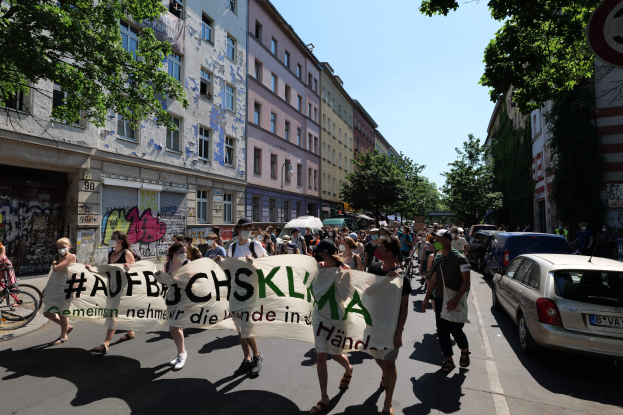 Eine Gruppe von Menschen marschiert auf einer Straße in Berlin, wobei sie ein Banner halten, mit parkenden Autos auf der rechten Seite, einem Fahrradfahrer auf der linken Seite und Gebäuden, Bäumen und einem Schild im Hintergrund unter einem sichtbaren Himmel.