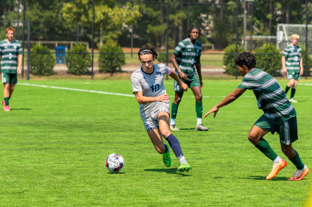 Junge Männer in Sportuniformen spielen Fußball auf einem grünen Feld mit Bäumen, Maschendrahtzaun und einem Torpfosten.
