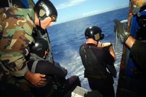 Drei Männer in Neoprenanzügen und Helmen sitzen in einem Hubschrauber, einer hält ein Objekt, mit dem Ozean und dem Himmel im Hintergrund.