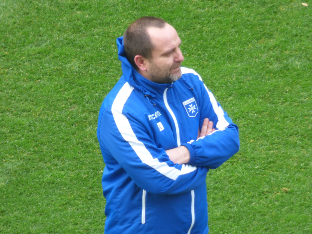 Ein Mann in einer blauen Jacke steht selbstbewusst auf einem grünen Feld und sieht aus wie ein Trainer.