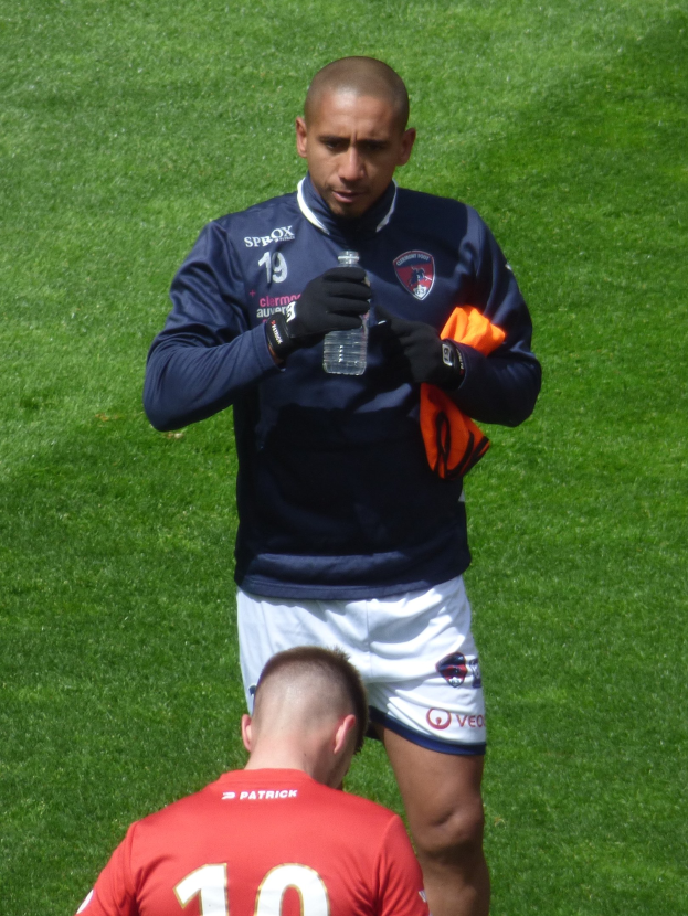 Ein professioneller Athlet steht auf einem grünen Feld und hält in einer Hand eine Flasche und in der anderen ein oranges Tuch.