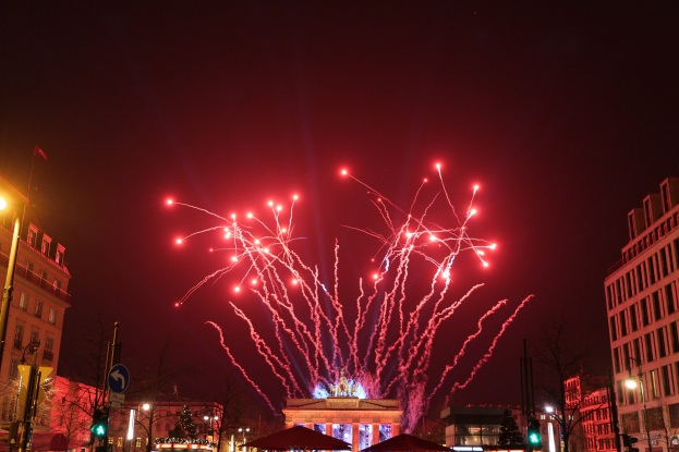 Eine belebte Stadtstraße bei Nacht während des Neujahrsfeier in Berlin, mit Gebäuden, Bäumen, Laternen, Verkehrszeichen, Zeltplanen, Menschen und einem prächtigen Feuerwerk am Himmel.